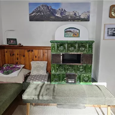 A cozy living room with a traditional tiled stove in green. Pictures of mountain landscapes hang on the wall, and there is a comfortable seating area.