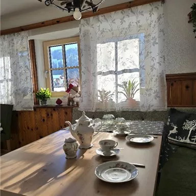 A cozy dining area with a wooden table, lovingly set. Bright curtains and some plants can be seen in the windows.