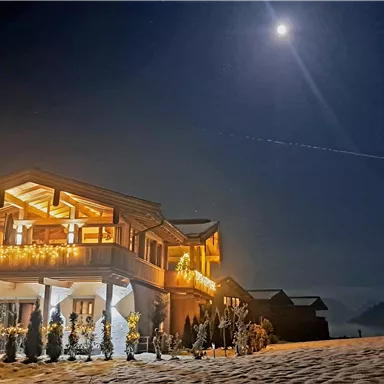 A cozy house at night, illuminated by warm lights. The brightly lit moon is in the sky, creating a tranquil atmosphere.