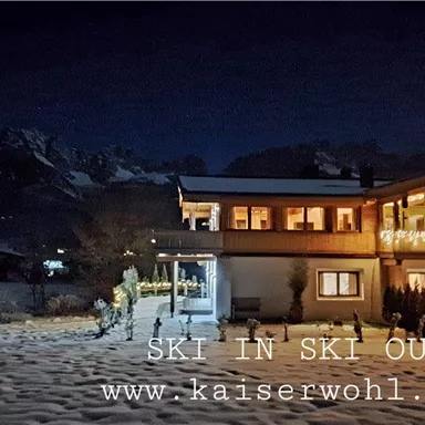A cozy chalet in the snow at night. In the background, mountain peaks can be seen, while the lights of the house shine.