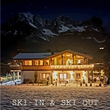 A cozy mountain cabin in a snow-covered landscape at night. In the background, the impressive mountains of the Wilder Kaiser can be seen.