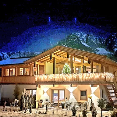 A beautiful wooden house at night, surrounded by snow-covered mountains. The building is festively illuminated and radiates a cozy atmosphere.