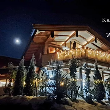 A beautiful chalet, festively decorated with lights, under a clear night sky. The moon shines over the Kaiserwohl chalet in a Christmas magic.