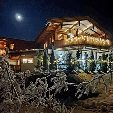 A winter house with festive lighting under a clear night sky. The snow surrounds the building and the moon shines brightly.