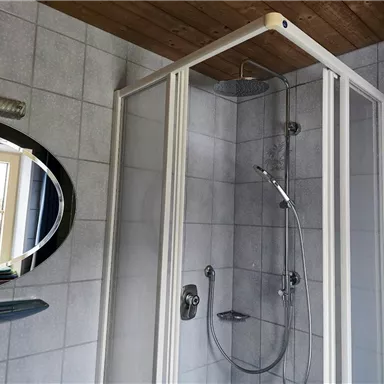 A modern shower cabin with glass hinged doors is located in a tiled bathroom. A round mirror and a practical shelf are also visible.