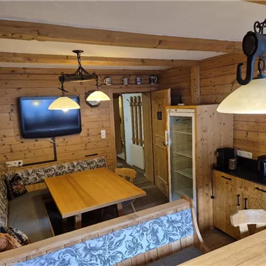 Ein gemütlicher Raum mit Holzwänden, einem Tisch und einer Sitzecke. An der Wand ist ein Fernseher und ein Kühlschrank steht in der Ecke.