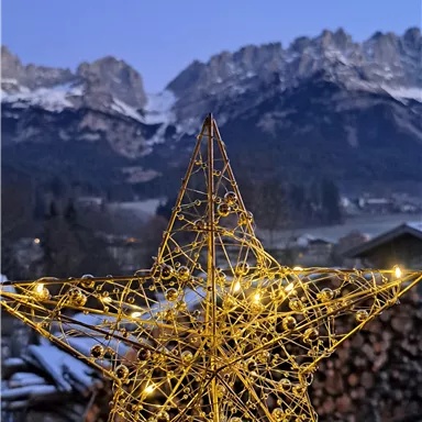 A glowing Christmas star made of wire lies on a table. In the background, snow-covered mountains and a clear, blue sky can be seen.