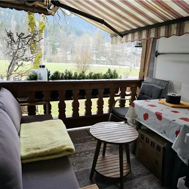 A cozy balcony with a couch, a small table, and a tablecloth with red dots. In the background, trees and a mountain landscape are visible.