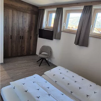 A modern bedroom with two beds and a television. The walls are designed in warm wooden tones and the windows let in plenty of daylight.