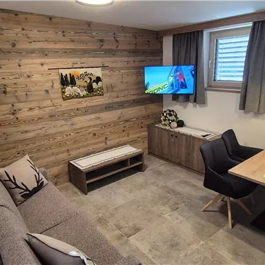 A cozy living space with wooden walls and a television. A dining table with black chairs and a sofa corner invites relaxation.