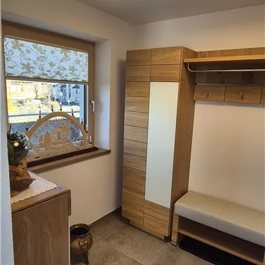 A cozy room with a small window and a modern wooden wardrobe. The floor is covered with a carpet, and there is a seating area.