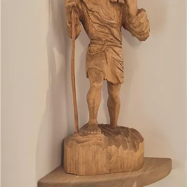 A wooden sculpture of a man with a child on his shoulder. The man is holding a walking stick and standing on a small shelf.