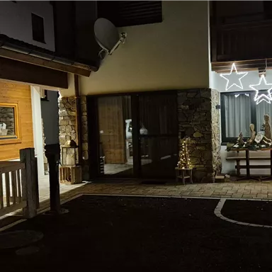 A festively decorated entrance area of a house with a Christmas tree and lights. The surroundings are illuminated at night, creating a cozy atmosphere.