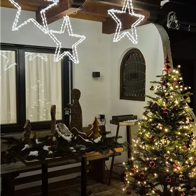 A festively decorated terrace with a lit Christmas tree and glowing stars. In the background, windows with warm light can be seen.
