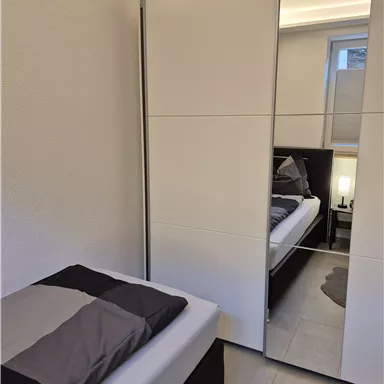 A modern bedroom with a single bed and a large mirror wardrobe. The walls are bright and the floor is tiled.