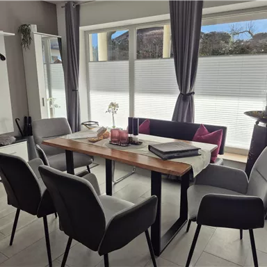 A modern dining area with a wooden table and four gray chairs. Large windows allow plenty of light in and offer a view of the outside.