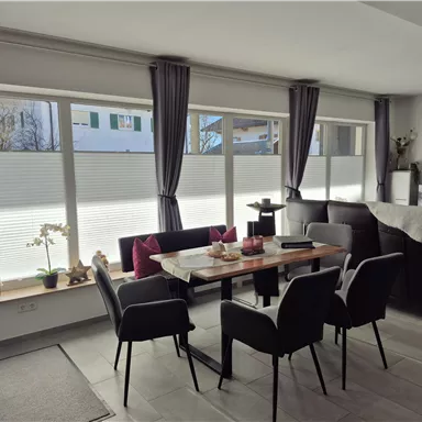 A modern dining room with a wooden table and black chairs. Large windows with sunlight illuminating the room.