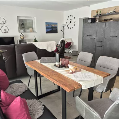 A modernly furnished dining area with a large wooden table and stylish chairs. The walls are neutral, and there are decorative elements like pictures and plants.