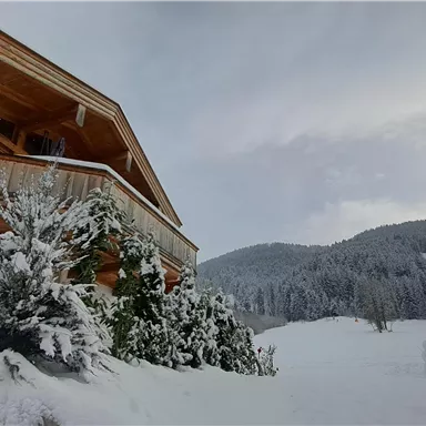 A cozy wooden house in a snowy winter landscape. In the background, there are wooded hills and a cloudy sky.