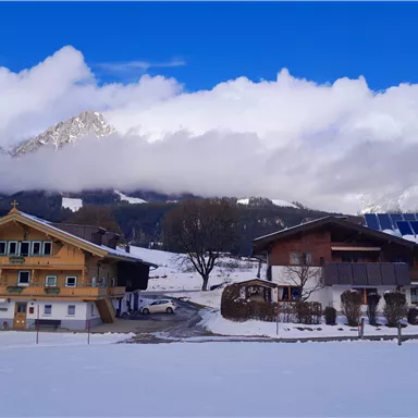 A picturesque winter landscape with snowy fields. Two traditional buildings stand before majestic mountains and a cloudy sky.