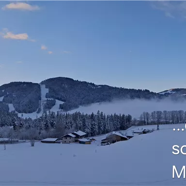 Eine verschneite Berglandschaft mit Hütten im Vordergrund. Der Himmel ist klar und es strahlt ein sanftes Morgenlicht.