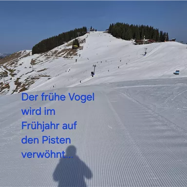 Eine schneebedeckte Piste mit vielen Skifahrern im Hintergrund. Die Sonne scheint auf die verschneite Landschaft.