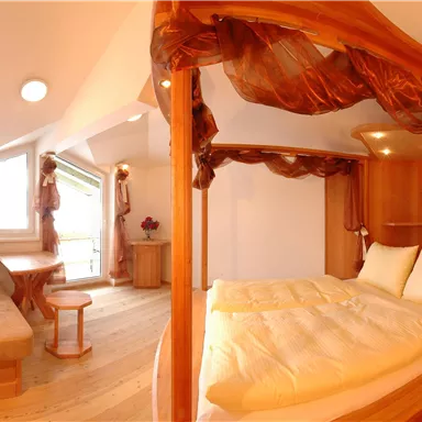 A cozy room with a wooden bed and light sheets. A loveseat and a table provide a pleasant atmosphere.