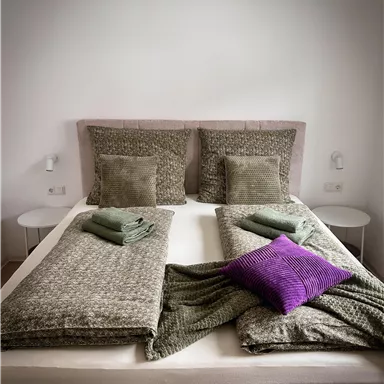 Ein modernes Bett mit zwei Kissen und verschiedenen Decken. Auf dem Bett liegen grüne Handtücher und ein lila Kissen.