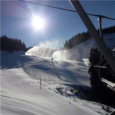 Eine schneebedeckte Piste unter einem klaren blauen Himmel. Im Hintergrund sprühen Schneekanonen frischen Schnee.