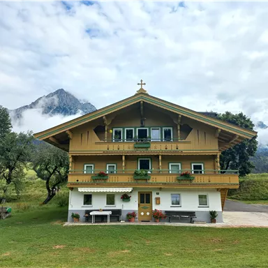 A beautifully situated house in Alpine style with a large balcony. In the background, mountains and a cloudy sky can be seen.