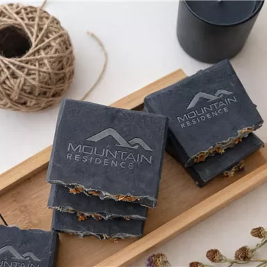 Ein stilvolles Arrangement von Seifen auf einem Holzbrett. Die Seifen sind mit dem Logo "Mountain Residence" graviert und umgeben von dekorativen Elementen wie Schnur und Kerze.