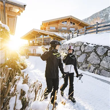 Two skiers are walking along a snowy path, surrounded by chalets. The sun is shining, creating a beautiful winter atmosphere.