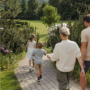 Eine Familie geht einen gepflasterten Weg entlang, umgeben von grünen Bäumen und Blumen. Im Hintergrund sind sanfte Berge zu sehen.