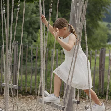 Ein Mädchen in einem weißen Kleid spielt auf einem Spielplatz. Sie überquert eine Wackelbrücke aus Seilen.