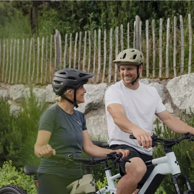 Ein Paar sitzt auf Fahrrädern und lächelt sich an. Im Hintergrund sind grüne Sträucher und ein Holzzaun zu sehen.