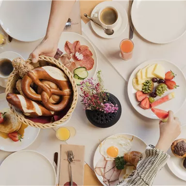 Ein bunter Frühstückstisch mit verschiedenen Speisen, darunter Brezeln, Obst und Gebäck. Es sind mehrere Teller und Getränke sichtbar, die eine gemütliche Atmosphäre schaffen.
