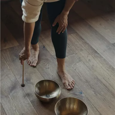 Eine Person steht barfuß auf einem Holzboden und spielt mit einem Holzschlägel auf zwei Schalen. Die Atmosphäre ist entspannend und ruhig.