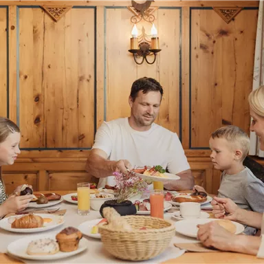 Eine Familie genießt ein Frühstückstisch mit verschiedenen Speisen. Die Atmosphäre ist gemütlich mit Holzwänden und fröhlichen Gesichtern.