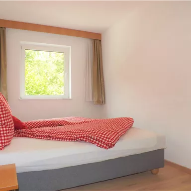 A cozy bedroom with a bed and a window. The bed linen has a red-and-white checked pattern and there is a bedside table next to it.