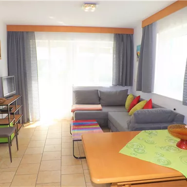 A bright living room area with a gray couch and colorful pillows. A table with a green tablecloth and windows that let in plenty of daylight.