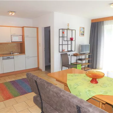 A modern apartment with an open kitchen and a cozy living area. The space is bright and welcoming, featuring a colorful carpet and a dining table.