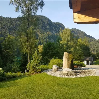 Eine ruhige Landschaft mit grünen Wiesen und hohen Bergen im Hintergrund. Steinformationen und Bäume schaffen eine harmonische Atmosphäre.
