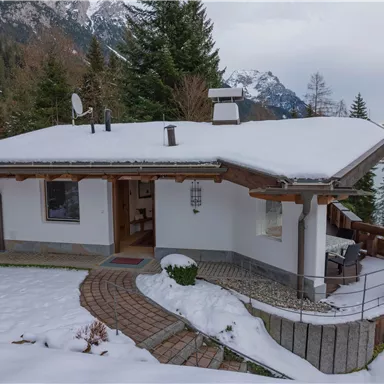 Ein gemütliches Haus im Schnee, umgeben von Bäumen und Bergen. Die verschneite Terrasse lädt zum Entspannen ein.