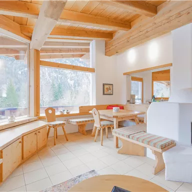 Ein einladendes Wohn- und Esszimmer mit großen Fenstern und Holzbalkendecke. Die gemütliche Einrichtung und der Blick auf die Natur schaffen eine warme Atmosphäre.