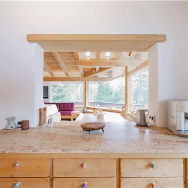 Eine helle Küche mit Holzdekor und einer großzügigen Theke. Im Hintergrund sind gemütliche Sitzmöglichkeiten und große Fenster mit Blick auf die Natur.