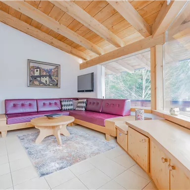 Ein gemütliches Wohnzimmer mit Holzdecken und modernen Möbeln. Große Fenster bieten einen Blick auf die Natur draußen.