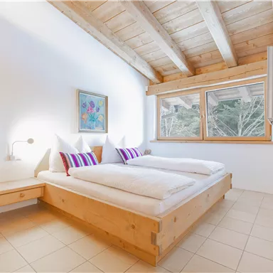 Ein gemütliches Schlafzimmer mit Holzdekor und großen Fenstern. Zwei Betten mit weißen Bettwäsche und bunten Kissen laden zum Entspannen ein.