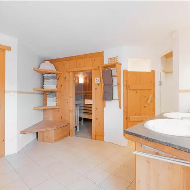 Ein modernes Badezimmer mit Holzverkleidung und zwei Waschbecken. Es gibt Regale mit Handtüchern und Zugang zu einem weiteren Raum.