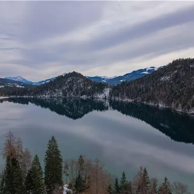 Ein ruhiger See umgeben von schneebedeckten Bergen und Tannenbäumen. Der Himmel ist bewölkt und das Wasser spiegelt die Landschaft wider.