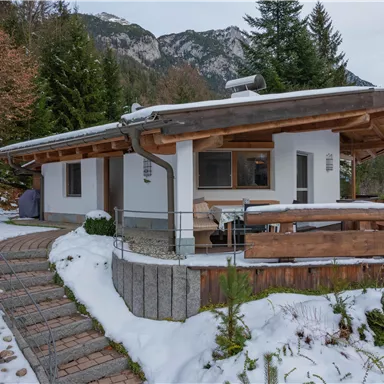 Ein gemütliches Ferienhaus im Schnee, umgeben von Bäumen und Bergen. Der Eingang ist mit einem gepflasterten Weg und einer Holzterrasse gestaltet.